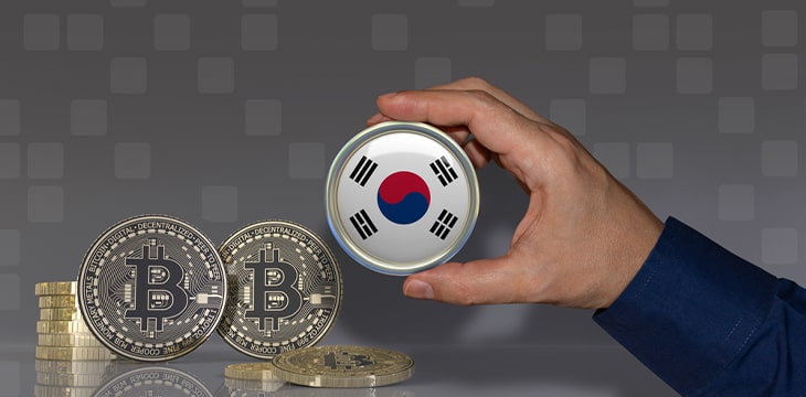 A hand holding a badge with the South Korean flag in front of some bitcoins. Crypto currency concept