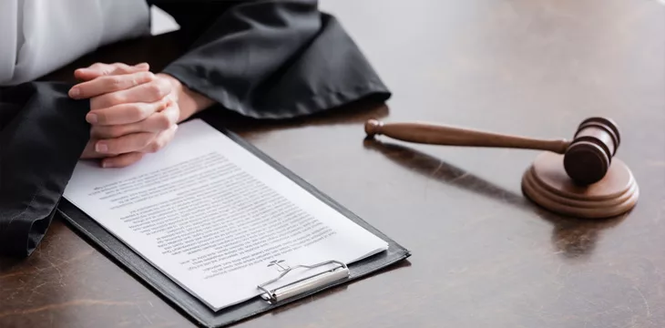 Partial view of prosecutor with clenched hands near lawsuit and gavel on wooden desk